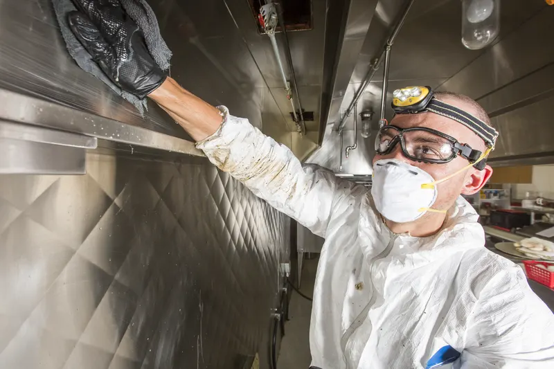Technician in gloves reaching up into the plenum of a stainless steel kitchen hood with the filters removed