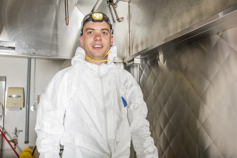 Technician in coveralls and a respirator cleaning a stainless commercial kitchen hood above a cook line