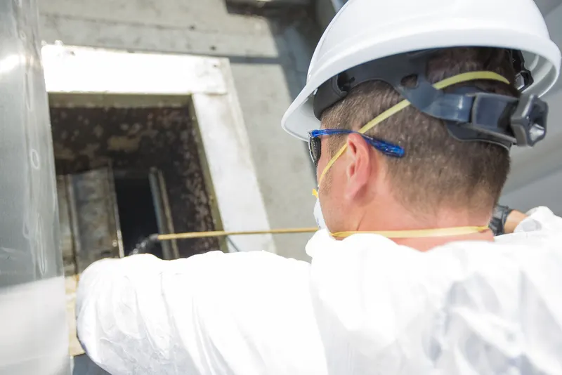 Technician reaching into an open access panel on a commercial kitchen exhaust duct above a ceiling